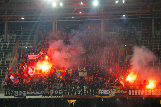 Eine große Menge Menschen in einem Stadion hält Fahnen und Banner, mit Leuchtraketen, unter einer Decke mit Deckenleuchten und Metallrahmen.