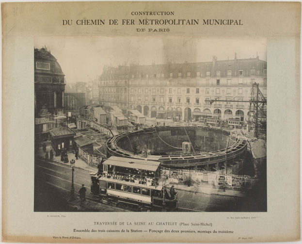 Ein altes Schwarz-Weiß-Foto einer Stadtstraße mit einem Zug auf den Schienen, umgeben von Gebäuden, Polen und Menschen, mit der Aufschrift "Construction du Chemin de Fer Metropolitain Municipal de Paris" oben.