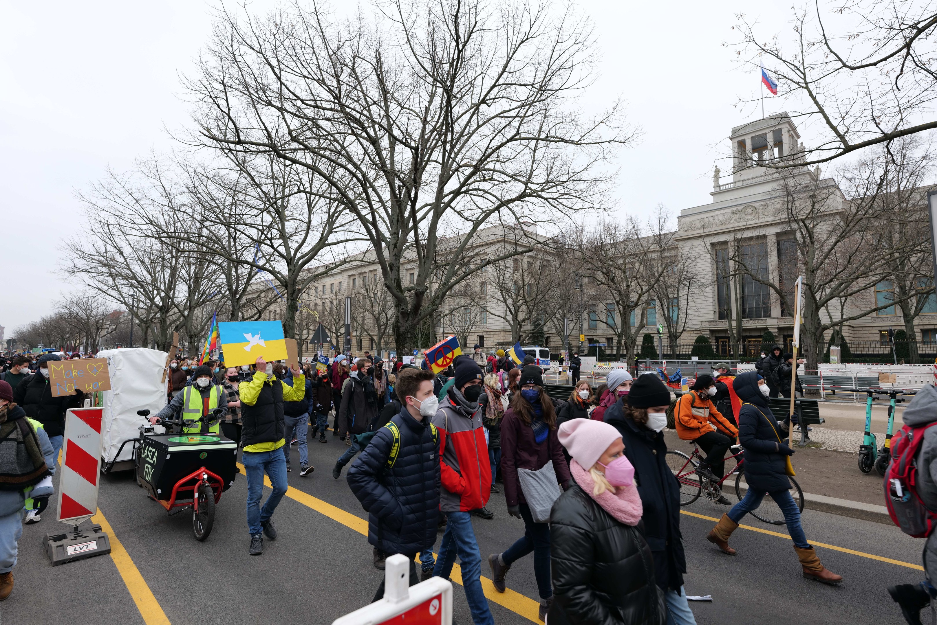 Eine große Gruppe von Menschen marschiert in Washington, D.C. im Protest die Straße entlang, mit Schildern und Bannern, während einige Fahrräder fahren, unter einem klaren blauen Himmel.