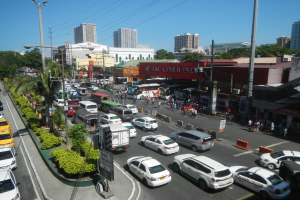 Eine belebte Stadtstraße mit Autos, Bussen, Fußgängern, Bäumen, Straßenlaternen, Strommasten und Gebäuden unter einem klaren blauen Himmel.