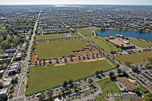 Luftaufnahme einer Stadt mit Gebäuden, Bäumen, Gras, Straßen mit Fahrzeugen, Wasser und bewölktem Himmel, die einen großen zentralen Platz zeigt, der als Park geplant ist.