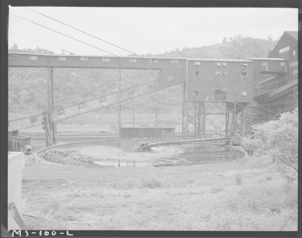 Ein Schwarz-Weiß-Foto eines Kohlebergwerks mit einer Brücke, Pfählen, Drähten, Gebäuden, Bäumen, Pflanzen, Gras und Wasser im Vordergrund und einem Himmel im Hintergrund, mit Text am unteren Rand.