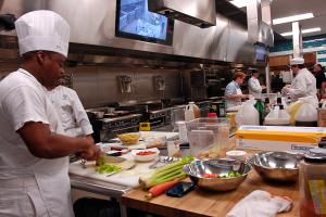 Eine Gruppe von Köchen in einer Küche beim Zubereiten von Essen auf einem Schneidbrett, mit einem Koch im Vordergrund in einer weißen Schürze und Mütze, der ein Messer hält und von verschiedenen Utensilien, Schüsseln, Gemüse, einem Mobiltelefon, Flaschen und anderen Gegenständen umgeben ist, mit einem Fernseher und Deckenleuchten im Hintergrund.