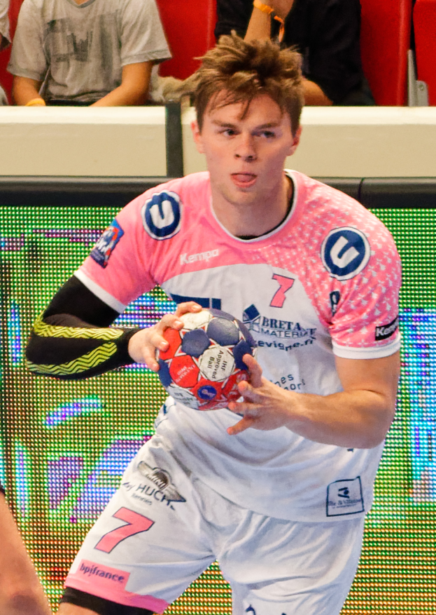 Ein Mann in einem pinken Hemd hält einen Fußball, mit einer anderen Person Hand auf der linken Seite und ein paar Menschen im Hintergrund auf Stühlen sitzend.