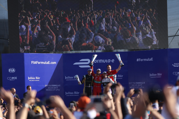 Gruppe von Menschen auf einem Formel-E-Podium in Mexiko-Stadt mit Pokale, Sponsor-Boards und einem Bildschirm im Hintergrund.