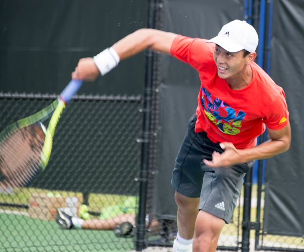 Ein Mann in rotem Hemd und weißer Mütze spielt Tennis und hält einen Schläger, mit einem Netz und durchscheinendem Hintergrund; eine weitere Person sitzt in der Nähe auf dem Boden mit einer Box und Bällen.