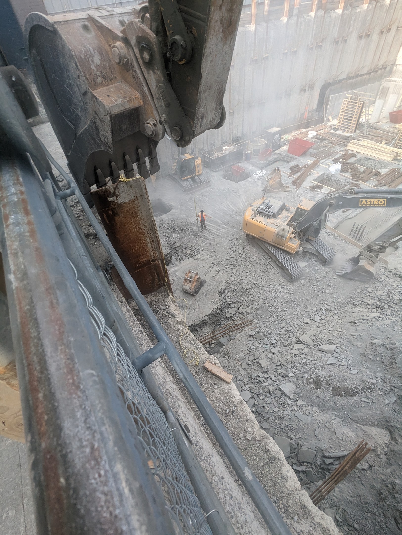 Baustelle mit einem Bagger, der ein Gebäude abbricht, Fahrzeuge, Menschen und ein Metallobjekt links.