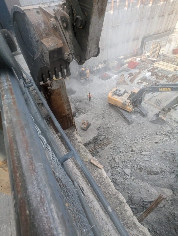 Baustelle mit einem Bagger, der ein Gebäude abbricht, Fahrzeuge, Menschen und ein Metallobjekt links.