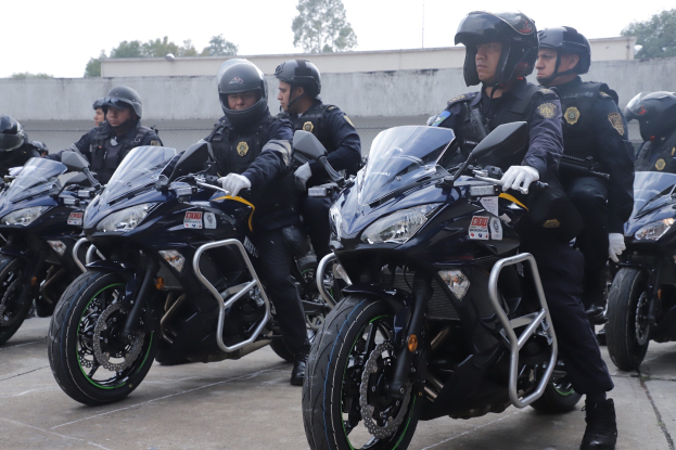 Polizeibeamte in schwarzen Uniformen, Helmen und Handschuhen auf Motorrädern in einer Parkanlage mit einer Wand, Bäumen und einem klaren blauen Himmel im Hintergrund.