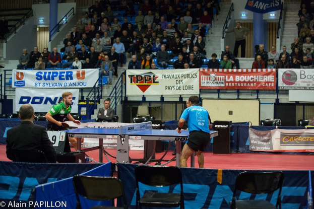 Zwei Männer spielen Tischtennis drinnen, mit mehreren Zuschauern, die stehen und sitzen, einigen leeren Stühlen und sichtbaren Werbeplakaten, mit Text in der linken unteren Ecke.