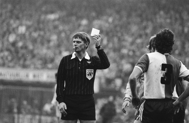 Schwarzes und weißes Bild eines Schiedsrichters mit roter Karte vor einer Menge in Fußballtrikots und einem Zaun im Hintergrund.