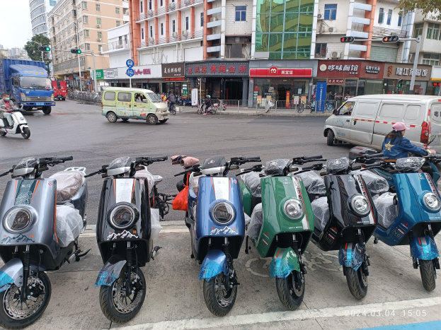 Eine Reihe von elektrischen Rollern, die auf einer Straße geparkt sind, mit einer Person in einer Möppe davor, vor dem Hintergrund von Gebäuden, Verkehrsampeln, Bäumen und einem klaren blauen Himmel.