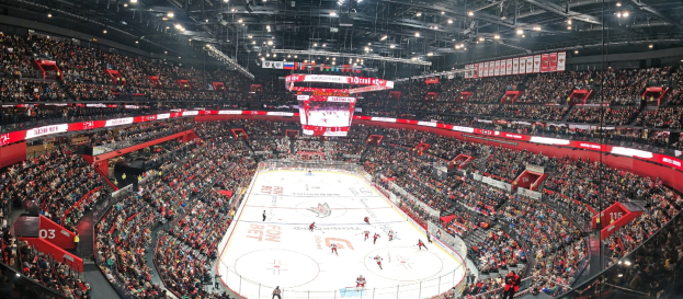 Eine große, hell erleuchtete Arena mit jubelnden Zuschauern, die ein Hockey-Spiel beobachten, mit einem zentralen Bildschirm über der Eisfläche.