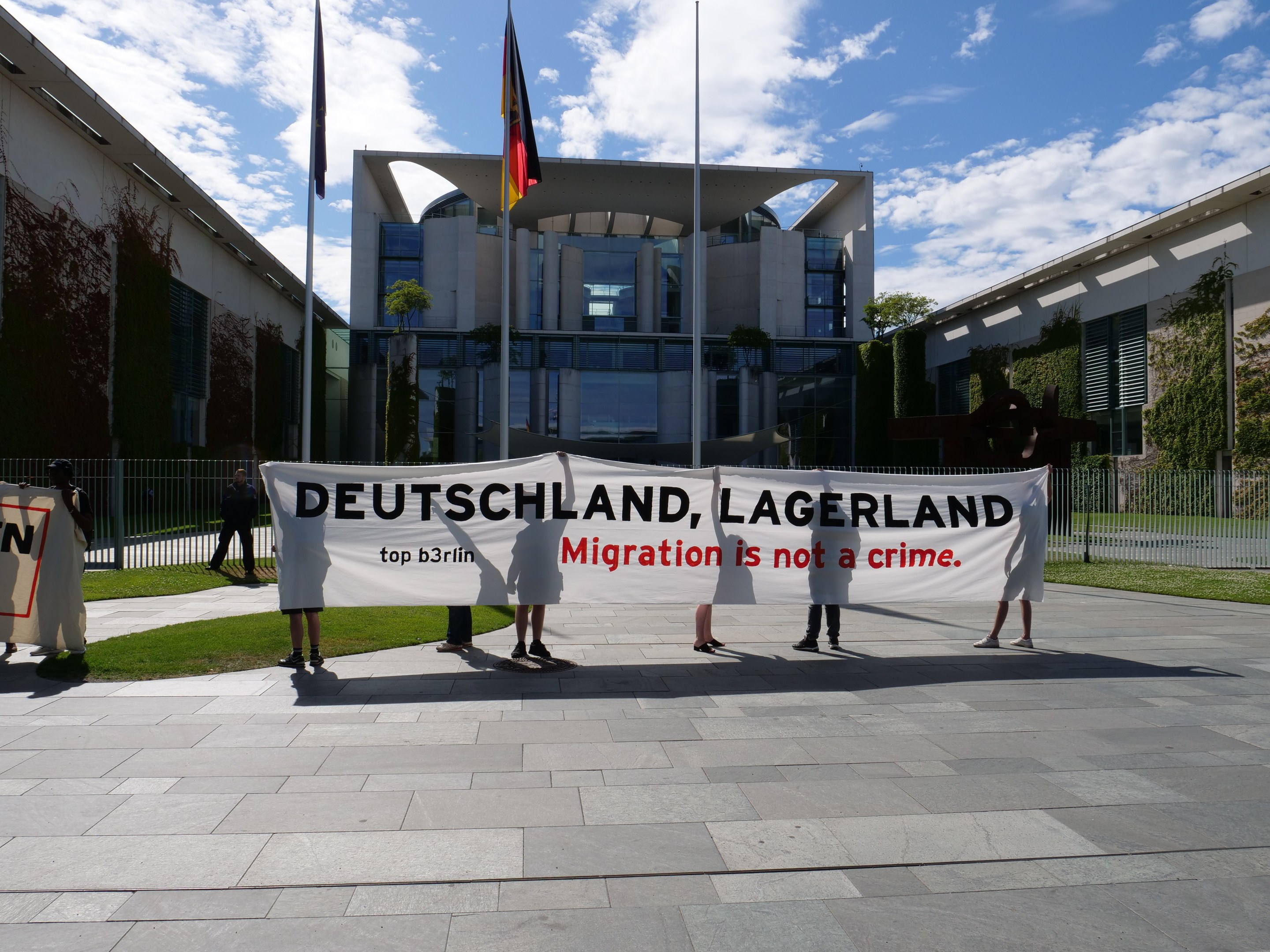 Eine Gruppe von Menschen, die ein Transparent mit der Aufschrift "Deutschland, Lagerland Migration ist kein Verbrechen" vor einem umzäunten Gebäude mit Bäumen und Fahnen halten.