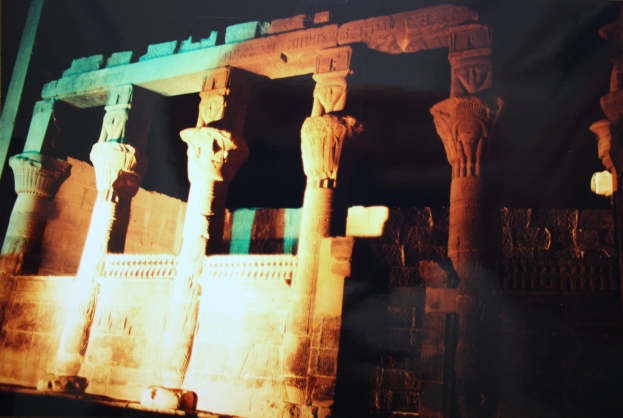 Ein altes Tempel mit Säulen und verzierten Wänden, beleuchtet von Licht, das hindurchscheint, vor einem dunklen Hintergrund.