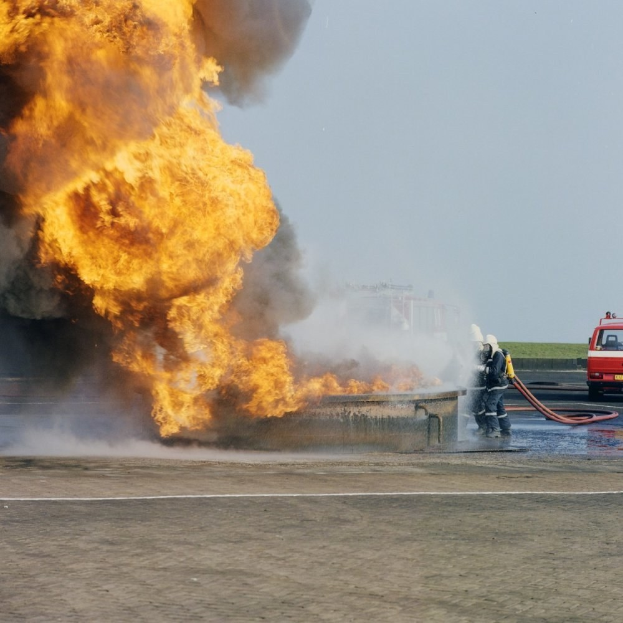 Feuerwehrlöschfahrzeug steht in Flammen an der Straßenseite mit zwei Helmen tragenden Personen, die Stäbe halten, einem Fahrzeug im Hintergrund und dem Himmel.