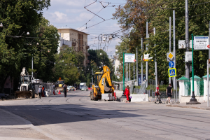 Eine Stadtstraße mit einer Baustelle in der Mitte, mit Fahrzeugen, Fußgängern, einem Radfahrer, Verkehrskegeln, Pfählen, Schildern, Strommasten mit Drähten, Bäumen, Gebäuden mit Fenstern und einem bewölkten Himmel im Hintergrund.
