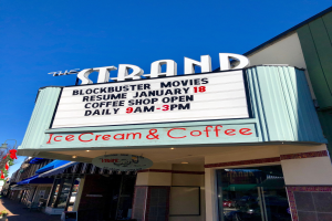 The Strand Blockbuster Movies Resume January 18 Coffee Shop building with a name board, a street pole, a group of trees, some plants in pots, and a cloudy sky.