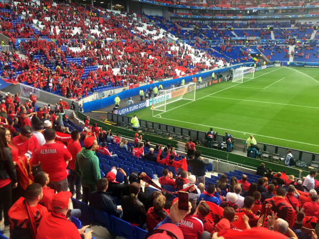 Ein volles Stadion mit Zuschauern, einige sitzen, andere stehen, die ein Fußballspiel beobachten, mit Bannern, einem Tor und Deckenbeleuchtung.