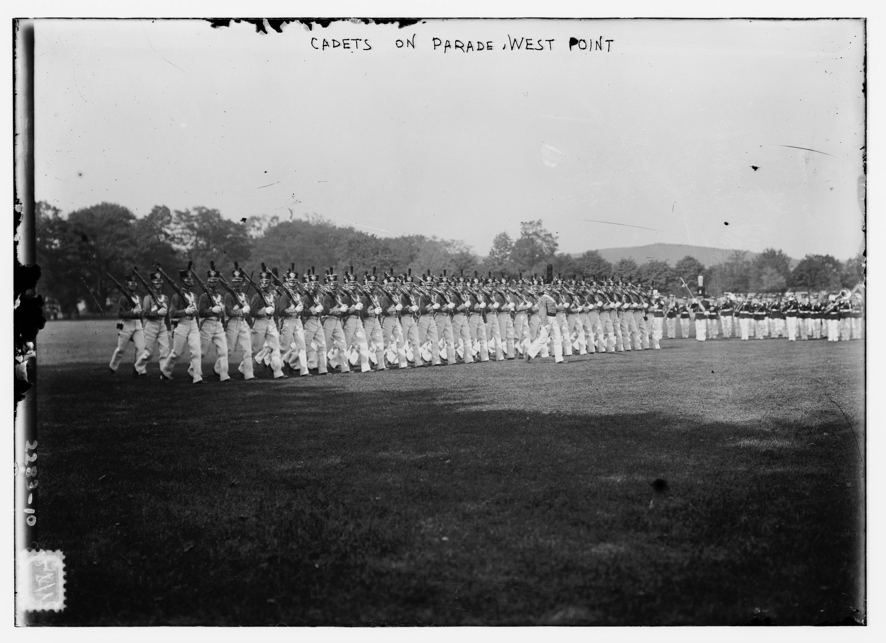 Gruppe von Menschen auf dem Boden stehend, einige halten Gewehre, mit Bäumen, Hügeln und einem bewölkten Himmel im Hintergrund, beschriftet "cadets on parade west point" oben.