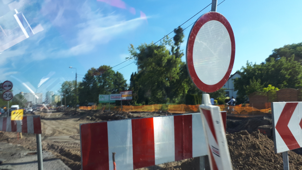 Baustelle mit einem roten und weißen Schild am Straßenrand, umgeben von Bäumen, Straßenlaternen, einem Verkehrsschild, Gebäuden und einem klaren blauen Himmel.