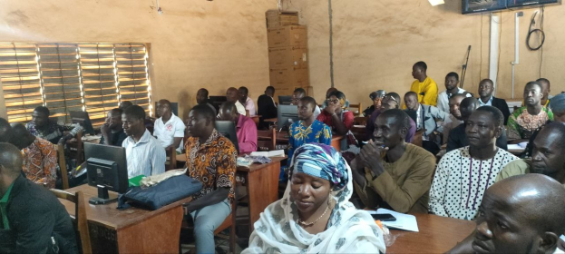 A group of people sits at desks in a classroom with monitors, bags, books, and papers on the tables, a window on the left, and cardboard boxes and a television on the wall in the background, attending a technology training session.