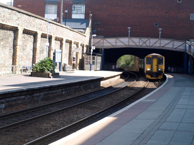 Ein Zug fährt auf den Schienen mit Plattformen auf beiden Seiten, einer Hauswand links, Straßenlaternen, Pflanzen, Bänken und einer Kabine davor, einer Brücke über die Plattformen und Gebäuden und Masten im Hintergrund.