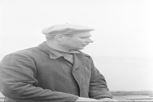 Ein altes Schwarz-Weiß-Foto eines Mannes in einer Jacke und einer Mütze, der nach rechts schaut und eine Uhr am Handgelenk trägt.