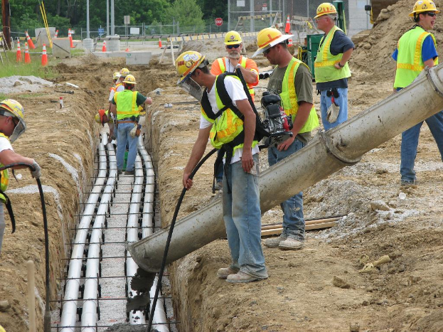 Eine Baustelle mit Arbeitern in Jacken und Helmen, Absperrbaken, Zaun, B√§ume, Masten, Erde und Rohrleitungen im Hintergrund.