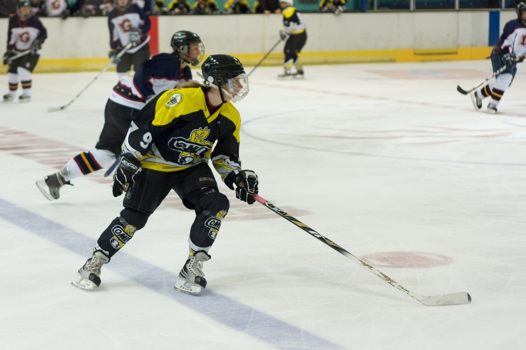 Menschen beim Eisstockschießen mit Eishockeyschlägern und Helmen, mit Zuschauern und einer Wand im Hintergrund.
