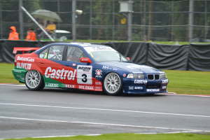 Ein Auto fährt auf einer Rennstrecke mit Gras auf beiden Seiten, mit Menschen in orangefarbenen Jacken und Schirmen hinter einem Maschendrahtzaun mit schwarzem Stoff und Bäumen im Hintergrund.