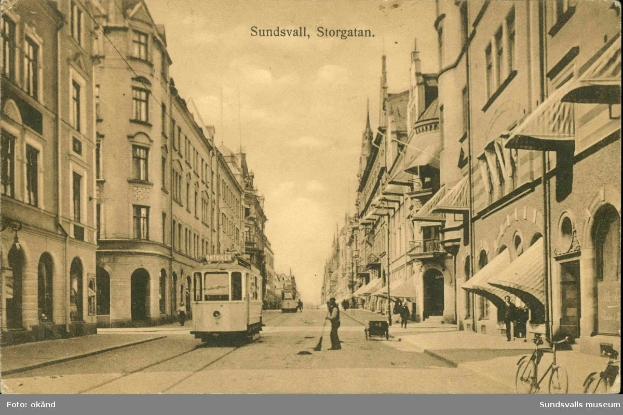Ein altes Foto einer Stadtstraße mit einer Tram, Gebäuden mit Fenstern, Menschen, die spazieren gehen, Fahrrädern und einem bewölkten Himmel, mit Text oben und unten auf dem Bild.