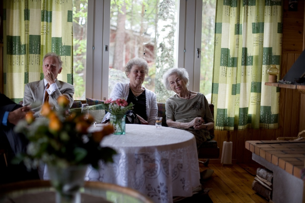 Eine Gruppe älterer Menschen sitzt auf einem Sofa mit einem Tisch davor, auf dem eine Blumenvase steht, und einer Glastür mit Vorhängen dahinter.