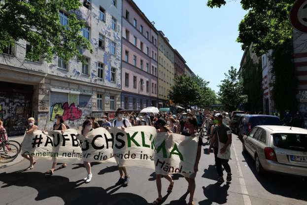 Menschen mit einem Transparent auf der Straße, mit parkenden Autos auf der rechten Seite und einem Radfahrer auf der linken Seite, vor Gebäuden und Bäumen während einer Demonstration in Berlin.