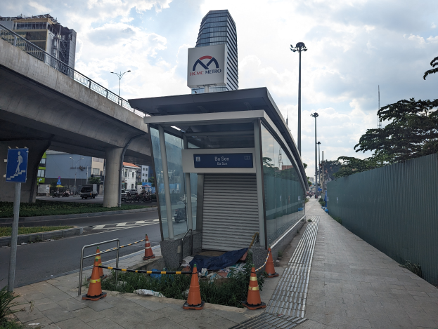 Eine Bushaltestelle am Straßenrand neben einem hohen Gebäude, umgeben von Absperrbaken, Pflanzen, Gras, einem Zaun, Fahrzeugen, einem Schild, Straßenlaternen, einer Brücke, Gebäuden mit Fenstern und einer bewölkten Himmel.