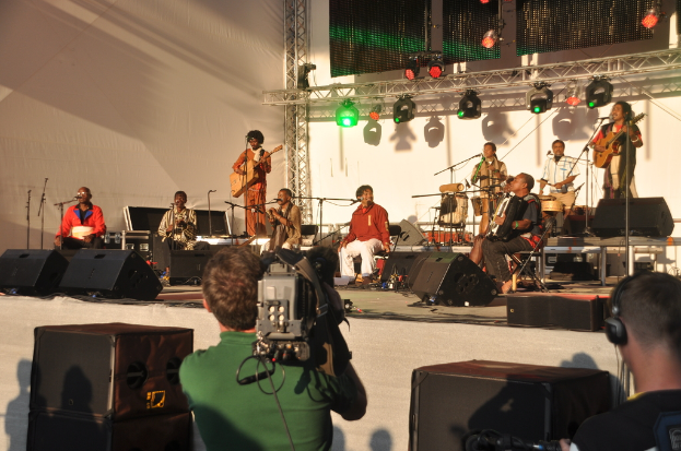 Ein musikalisches Konzert mit verschiedenen Musikern auf der Bühne, darunter Gitarren und Trommeln, unter rotem und grünem Bühnenlicht, mit Lautsprechern unten und zwei Personen, eine Aufnahme und die andere zuhörend.