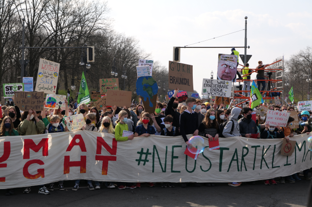 Eine große Gruppe von Menschen marschiert auf einer Straße, hält eine 'Menschenrechte'-Plakette und verschiedene Schilder, mit Bäumen, Laternenmasten und einem klaren blauen Himmel im Hintergrund.