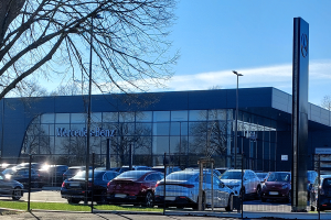Ein Mercedes-Autohaus mit parkenden Autos vorne, umgeben von einem Metallzaun, Gras, einem Schild, Straßenlaternen, einer Gruppe von Bäumen und einem bewölkten Himmel.