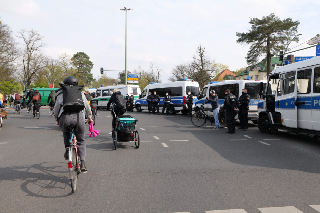 Eine Gruppe von Menschen, die auf Fahrrädern eine Straße entlangfahren, die von Polizeiwagen und anderen Fahrzeugen gesäumt ist, einige tragen Helme und Taschen, andere schieben Kinderwagen, mit Bäumen, Laternenmasten, Verkehrsampeln, Schildern, Gebäuden und einem bewölkten Himmel im Hintergrund.