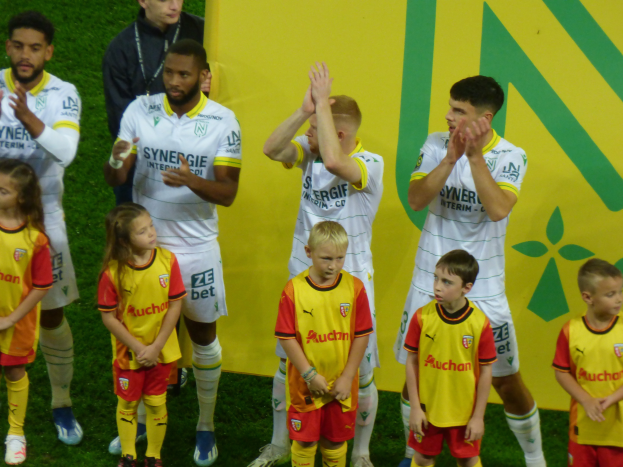 Eine Gruppe junger Menschen in Fußballtrikots steht auf einem Fußballfeld und klatscht feierlich mit einem "Ligue 1"-Schild im Hintergrund.