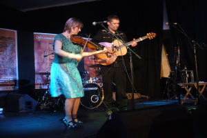 Ein Mann in schwarzem Hemd und Hose spielt Gitarre auf der Bühne, begleitet von einer Frau in einem blauen Kleid, die Geige spielt, mit verschiedenen Musikinstrumenten und Lautsprechern im Hintergrund.