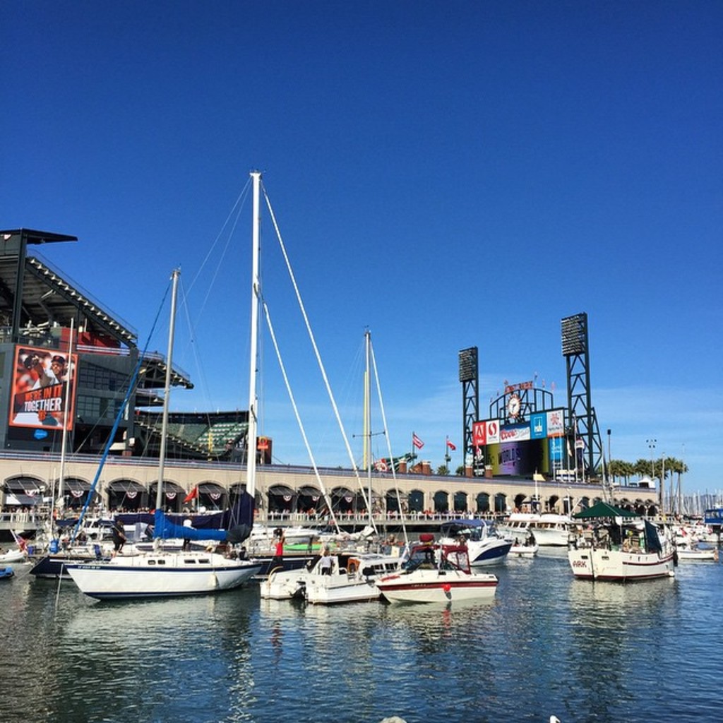 Viele Boote im Wasser im Vordergrund, mit einer Brücke mit mehreren Bögen, Fahnen, Bannern, Gebäuden, Pfählen, Bäumen und einem bewölkten Himmel im Hintergrund.
