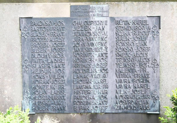 Ein Steinplakette an einer Wand mit eingraviertem Text, umgeben von Pflanzen am Boden, wahrscheinlich zur Erinnerung an Holocaust-Opfer.