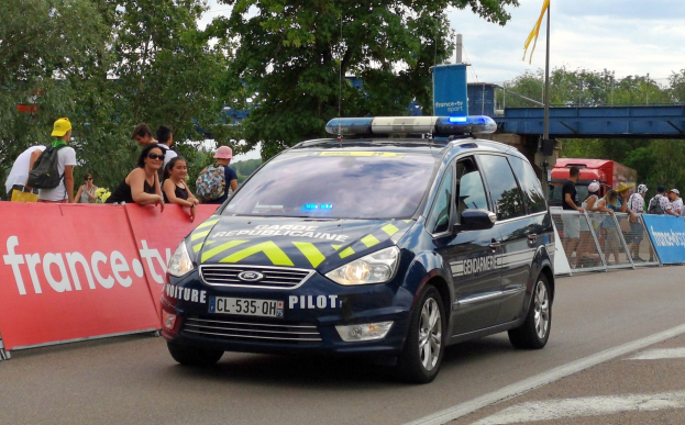 Polizeiauto fahrt an einer Menge mit Schildern, Gel├Ąndern, B├Ąumen, einer ├Überquerung, einer Flagge und einem bew├Âlktem Himmel im Hintergrund vorbei.