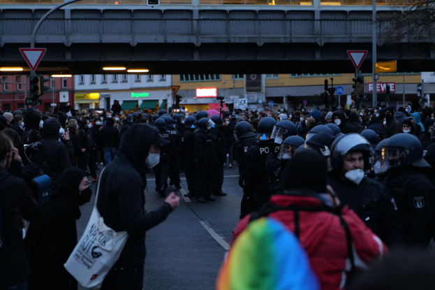 Große Gruppe von Menschen vor einer Reihe von Polizeibeamten, einige mit Helmen und Taschen oder Kameras, mit Gebäuden, Schildern, Laternenmasten, Verkehrszeichen, einer Brücke und einem Baum im Hintergrund.