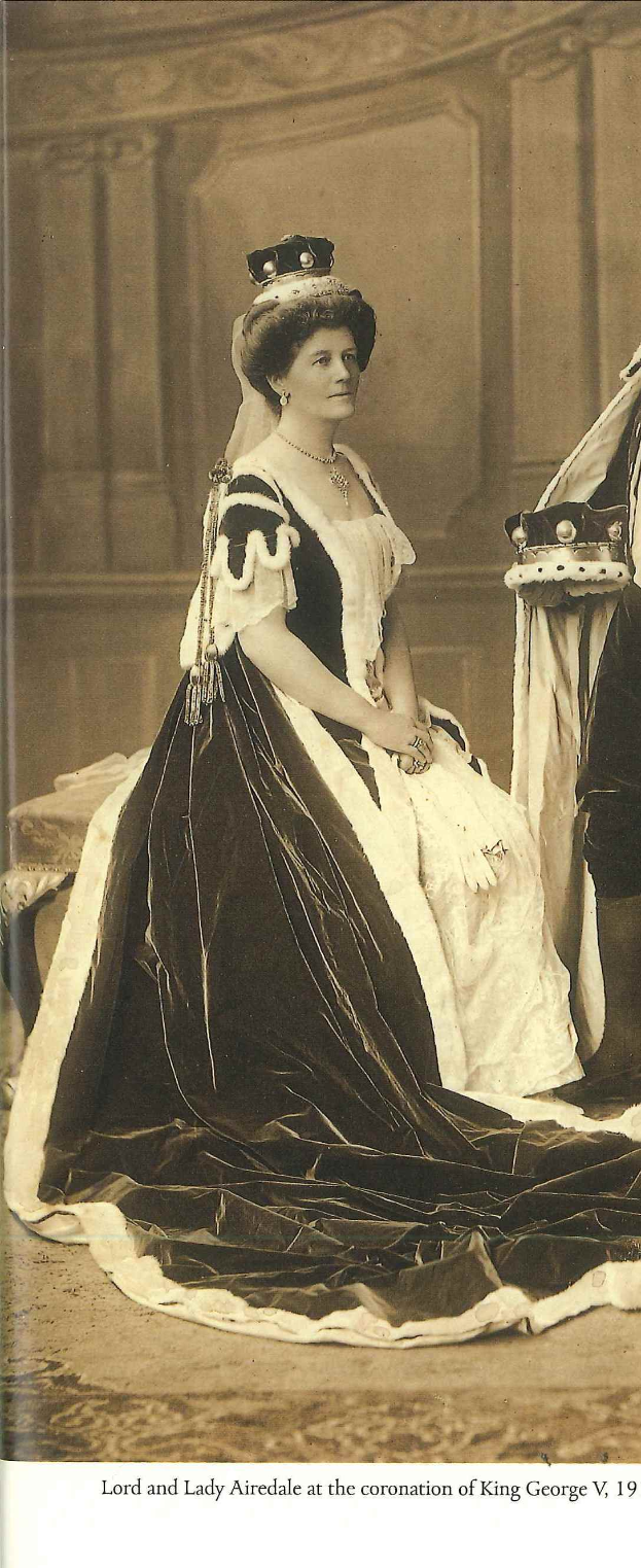 Lady Airedale, in einem schwarzen und weißen Kleid, steht vor einer Wand mit einem Objekt rechts daneben und Text unten im Bild.