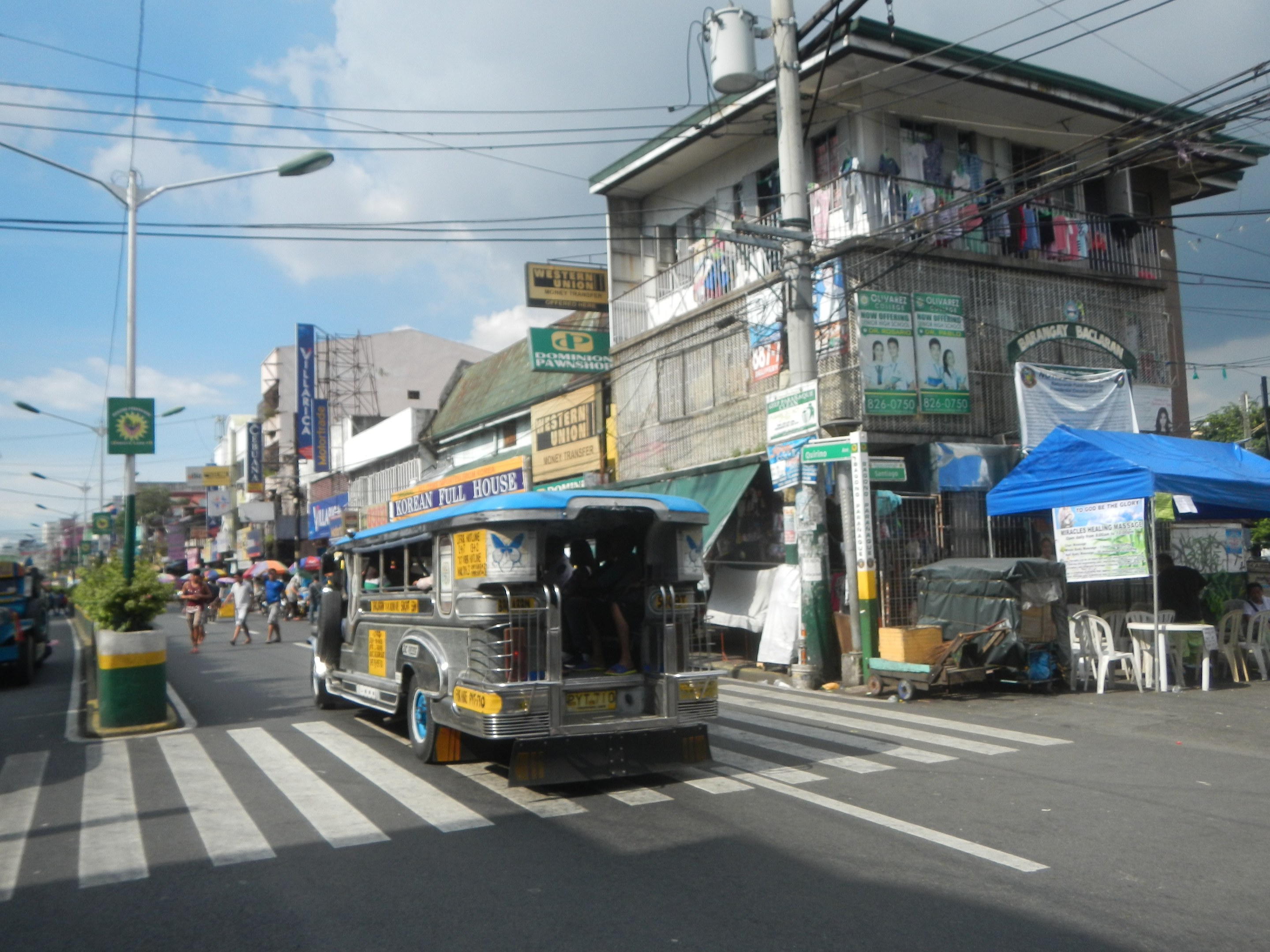 Eine Straßenansicht mit einem Bus, Gebäuden, Straßeninfrastruktur, Fahrzeugen, Fußgängern, Außensitzgelegenheiten, Grünfläche und einem bewölkten Himmel.