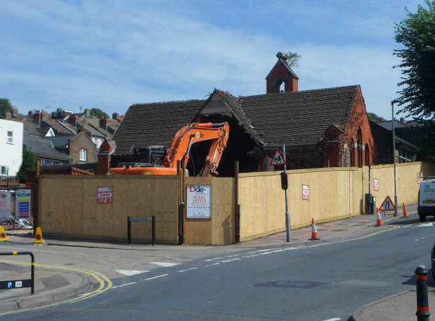Straße mit Fahrzeugen, Verkehrsleitkegeln, Pfosten, Schildern und einem Zaun; Bagger und abgerissenes Gebäude am Straßenrand; Gebäude, Bäume, Wolken und Himmel im Hintergrund.