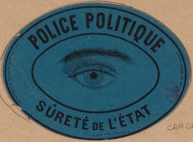 Ein brauner Karton mit einem blauen Aufkleber, auf dem "Polizei Politique S√Ľret√© de l'Etat" steht und ein Auge in der Mitte hat.