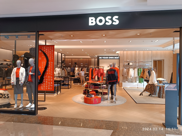 Das Innere eines Hugo Boss Stores in einem Einkaufszentrum, mit Schaufensterpuppen in Kleidung, Kleider an Bügeln, Tischen mit verschiedenen Gegenständen, beleuchteter Decke und einem Textschild oben.